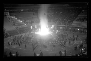 Schwarzes und weißes Foto eines vollen Stadions mit beleuchteten Stufen und Geländern, das eine zentrale Bühne mit einem hellen Feuer zeigt.