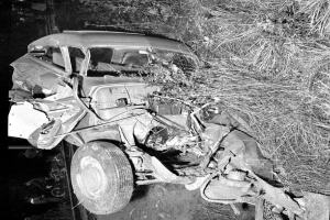 Ein Schwarz-Weiß-Foto eines schwer beschädigten Autos am Straßenrand mit einer eingedrückten Front und zersplitterten Fenstern, umgeben von verstreuten Gegenständen und einem Baum im Hintergrund.