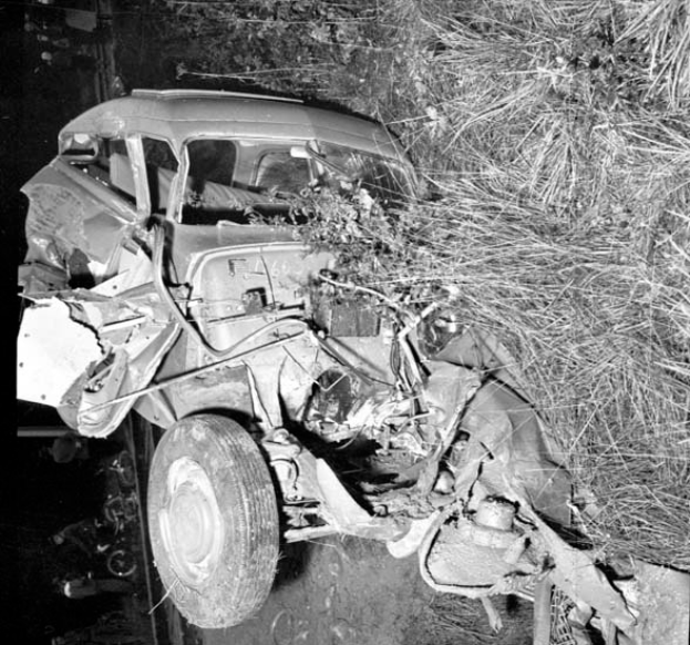 Ein Schwarz-Weiß-Foto eines schwer beschädigten Autos am Straßenrand mit einer eingedrückten Front und zersplitterten Fenstern, umgeben von verstreuten Gegenständen und einem Baum im Hintergrund.