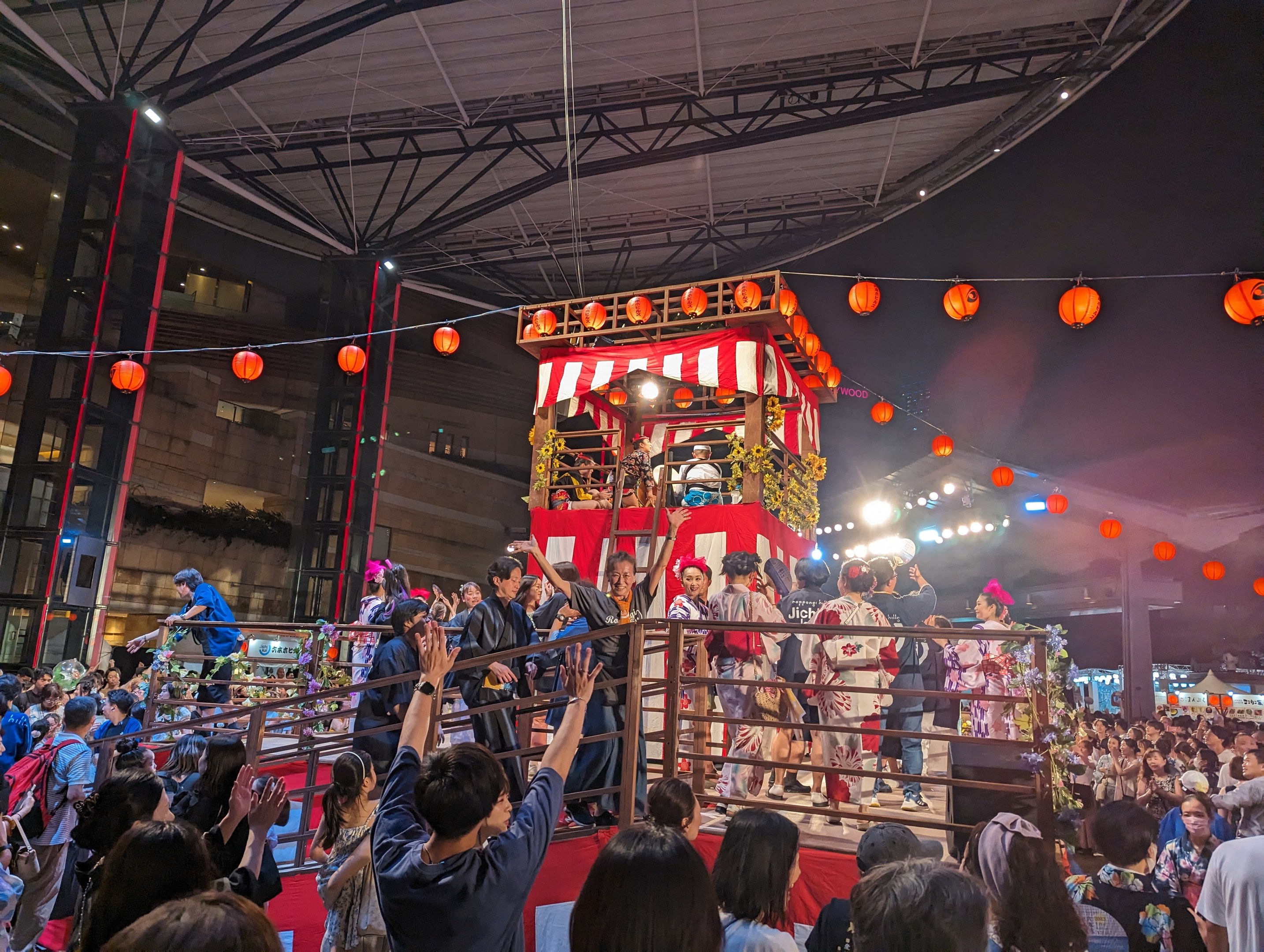 Menschenmenge auf einer beleuchteten Bühne mit Laternen, einige halten Kameras, umgeben von einem Geländer, mit Bäumen und einem klaren blauen Himmel im Hintergrund bei Nacht, möglicherweise während eines japanischen Festivals.