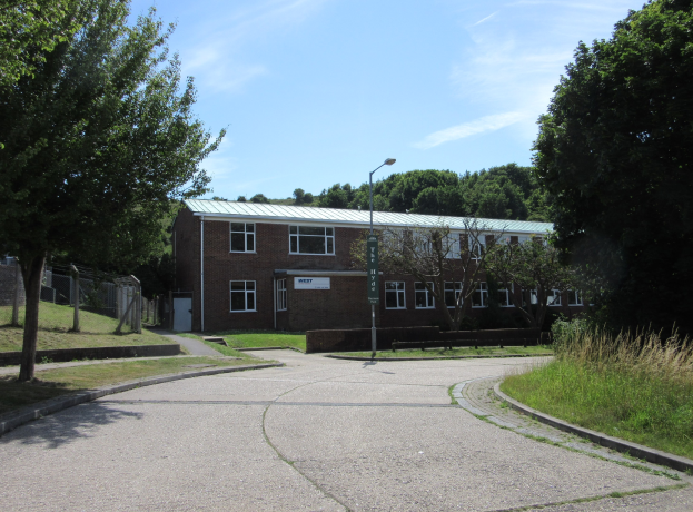 Ein Schulgebäude mit Fenstern, einem Straßenschild, einem Schild, einer Baumgruppe, Gras, einem Zaun und einer wolkenverhangenen Himmel, das in der Mitte eines Parks umgeben von saftigem Grün liegt.