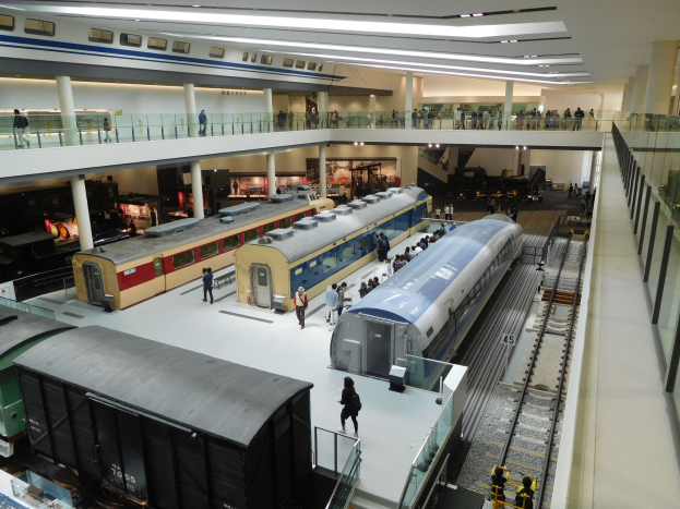 Eine großzügige Museumsausstellung mit mehreren Zügen in verschiedenen Größen und Farben, bei der Besucher die Ausstellungsstücke, Geländer, Deckenleuchten und tragenden Säulen betrachten.