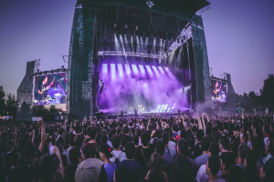 Große Menschenmenge auf einem Musikfestival mit Künstlern auf einer beleuchteten Bühne, umgeben von Bildschirmen, unter einem klaren blauen Himmel mit Bäumen und einem Gebäude im Hintergrund.