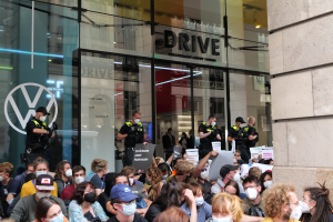 Große Gruppe von Menschen mit Masken und Schildern vor einem Glasgebäude mit Text, einige tragen Mützen und halten Kameras während einer Demonstration in Melbourne, Australien.