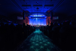 Ein großer Auditorium mit sitzenden Menschen, eine Bühne mit Musikern und eine Decke mit Lichtern und Fernsehern, was auf einen Konzertsaal für die Nationale Convention 2019 hindeutet.