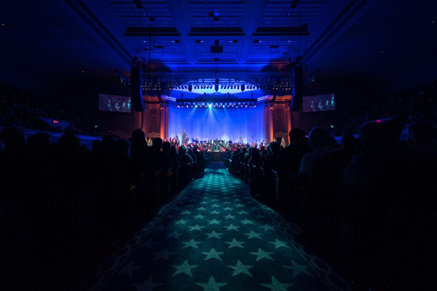 Ein großer Auditorium mit sitzenden Menschen, eine Bühne mit Musikern und eine Decke mit Lichtern und Fernsehern, was auf einen Konzertsaal für die Nationale Convention 2019 hindeutet.