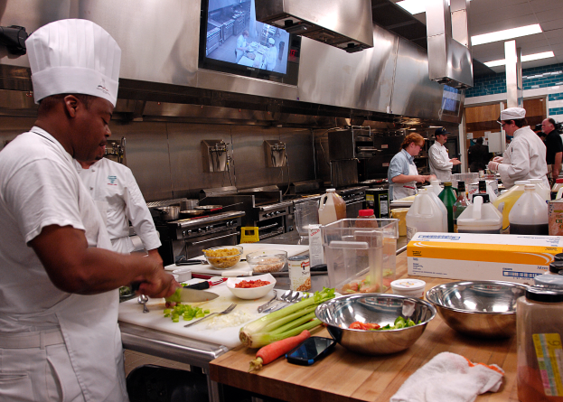 Eine Gruppe von Köchen in einer Küche, die Essen auf einem Schneidbrett zubereiten, mit einem Koch im Vordergrund in einer weißen Schürze und Mütze, der ein Messer hält, und umgeben von verschiedenen Utensilien, Schüsseln, Gemüse, einem Mobiltelefon, Flaschen und anderen Gegenständen, mit einem Fernseher und Deckenleuchten im Hintergrund.