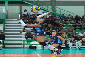 Eine weibliche Volleyballspielerin springt, um den Ball in der Luft zu treffen, mit zwei anderen Frauen im Vordergrund und Zuschauern im Hintergrund.