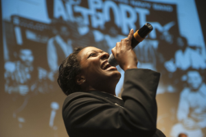 Eine Frau in einem schwarzen Kleid singt leidenschaftlich in ein Mikrofon auf der Bühne, ihre Augen geschlossen und ihr Gesichtsausdruck intensiv, mit Scheinwerfern, die ihr Gesicht beleuchten.