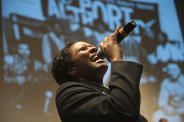 Eine Frau in einem schwarzen Kleid singt leidenschaftlich in ein Mikrofon auf der Bühne, ihre Augen geschlossen und ihr Gesichtsausdruck intensiv, mit Scheinwerfern, die ihr Gesicht beleuchten.