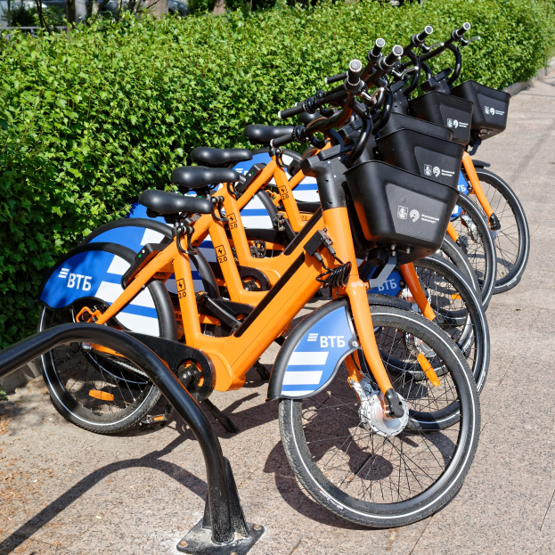 Eine Reihe von orange und blauen Fahrrädern, die nebeneinander auf der Seite einer Straße geparkt sind, mit Pflanzen und Bäumen im Vordergrund und einem Gebäude im Hintergrund.