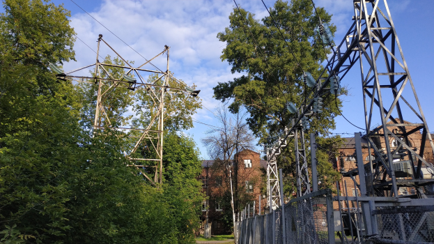 Gro├če Strommast mit mehreren Dr├Ąhten und Pfosten, die durch einen bewaldeten Bereich verlaufen, mit im Hintergrund sichtbaren Geb├Ąuden unter einem bew├Âlktem Himmel.