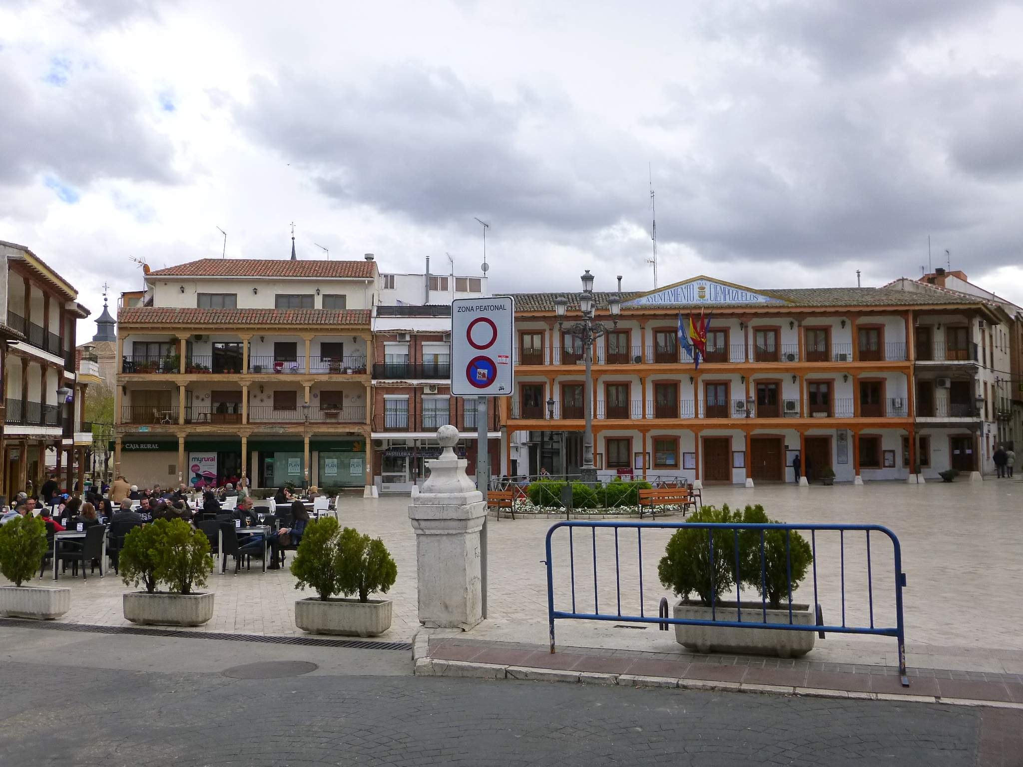 Ein belebter Stadtplatz mit sitzenden und stehenden Menschen, Topfpflanzen, Metallabsperrungen, einem Schild an einem Pfahl, Straßenlaternen mit Fahnen, umliegende Gebäude mit Fenstern und einem bewölkten Himmel.