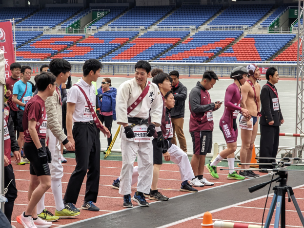 Gruppe von Menschen auf einer Bahn mit einer Kamera auf einem Stativ rechts, Hintergrund enthält Stühle, Geländer und eine Fahne.