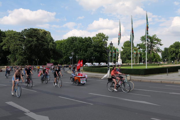 Eine Gruppe von Menschen, die auf Fahrrädern eine von Bäumen gesäumte Straße mit Laternenmasten und Fahnenmasten entlangfahren, während der Pariser Fahrradtour 2016, mit bewölktem Himmel und Fahrzeugen im Hintergrund.