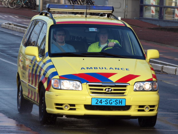 Ein gelber Krankenwagen mit zwei sichtbaren Insassen fährt an parkenden Fahrrädern auf einem Gehweg vorbei, mit einem Glasfrontgebäude im Hintergrund.