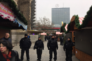 Polizisten in schwarzen Uniformen stehen vor einem Berliner Weihnachtsmarkt mit festlicher Dekoration, Gebäuden, Bäumen und einem klaren blauen Himmel im Hintergrund; ein Schild mit Text ist links sichtbar.