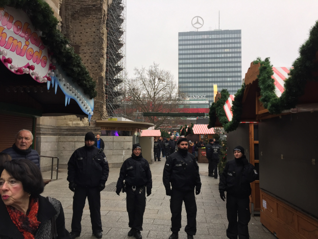Polizisten in schwarzen Uniformen stehen vor einem Berliner Weihnachtsmarkt mit festlicher Dekoration, Gebäuden, Bäumen und einem klaren blauen Himmel im Hintergrund; ein Schild mit Text ist links sichtbar.