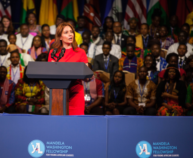 Eine Frau steht an einem Podium mit Mikrofonen, spricht zu einer Menge und hat im Hintergrund eine Tafel mit Text und Logos.