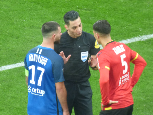 Schiedsrichter in schwarzem Hemd und Hose, der mit zwei Männern auf einem Fußballfeld spricht, einer in einem blauen und roten T-Shirt mit Text, beide tragen Brillen.