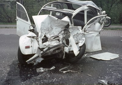 Schwerbeschädigte Autos am Straßenrand mit Frontalaufprall, Bäume und ein Gebäude im Hintergrund, weiteres Fahrzeug in der Ferne sichtbar.