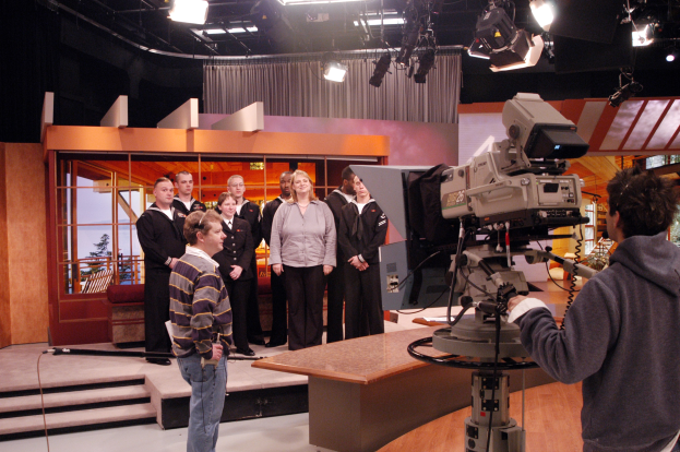 Eine Gruppe von Menschen in einem Fernsehstudio mit einer Person, die eine Kamera hält, auf der rechten Seite und Deckenleuchten im Hintergrund.