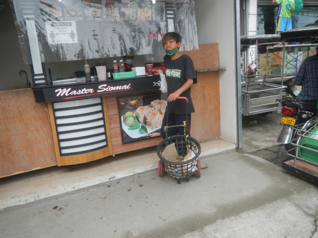 Ein junger Junge in einem schwarzen T-Shirt und einer Gesichtsmaske hält einen Einkaufswagen mit einem Korb, der vor einem Essensstand steht, mit zwei Personen auf einem Roller rechts daneben und einigen weiteren Personen im Hintergrund.