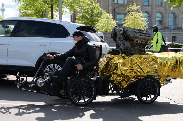 Ein Mann im Rollstuhl mit einem großen Motor am Rücken, umgeben von Fahrzeugen auf einer Straße mit Bäumen, Gebäuden und einem klaren blauen Himmel im Hintergrund, trägt eine schwarze Jacke und eine Mütze und hält ein Objekt.