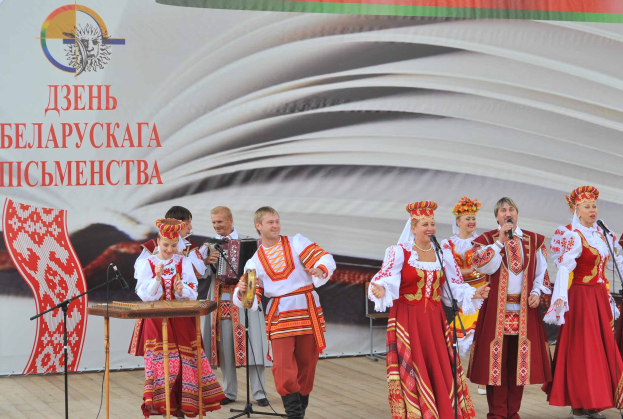 Eine Gruppe von Menschen in roter und weißer Kleidung performs auf der Bühne mit Mikrofonen und Instrumenten, mit einem "Ukrainischen Folklore-Festival"-Banner im Hintergrund.
