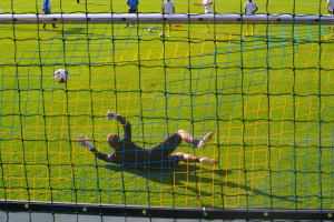 Ein Fussballtorhüter beim Abwehren eines Schusses vor dem Tor, mit einem Netz im Vordergrund und einigen Personen im Hintergrund, während mehrere Fussbälle auf dem Boden verstreut sind.