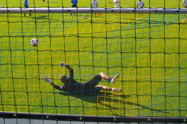 Ein Fussballtorhüter beim Abwehren eines Schusses vor dem Tor, mit einem Netz im Vordergrund und einigen Personen im Hintergrund, während mehrere Fussbälle auf dem Boden verstreut sind.