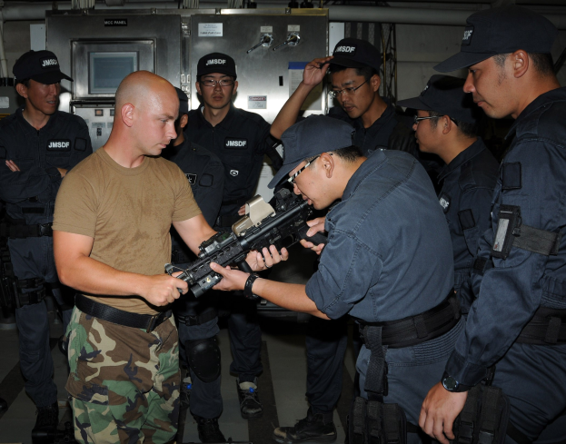 Eine Gruppe von Männern in Mützen mit Waffen, die in der Nähe von Industriegeräten und Rohren stehen.