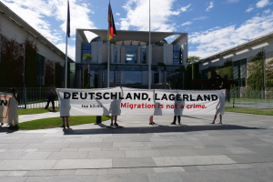 Eine Gruppe von Menschen hält ein Transparent mit der Aufschrift "Deutschland, Lagerland Migration ist kein Verbrechen" vor einem Gebäude mit Fenstern, umgeben von Gras, einem Metallzaun, Pflanzen, Bäumen und Fahnenmasten unter einem bewölkten Himmel.