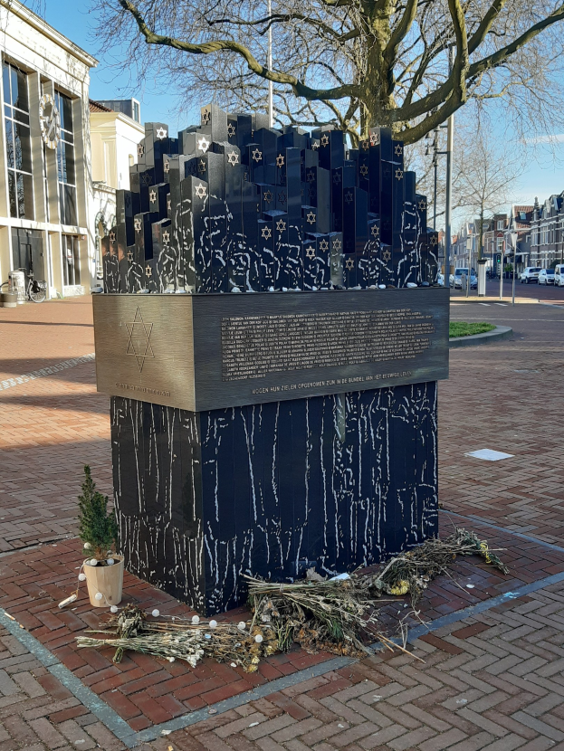 Denkmal für Holocaust-Opfer in einem Stadtplatz mit einem Baum davor, einem Topfgewächs auf dem Boden, verstreuten trockenen Blättern und umliegenden Gebäuden, Fahrzeugen, Pfählen und Bäumen.