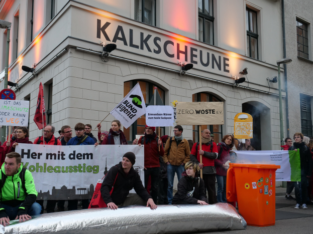Eine Gruppe von Menschen hält Schilder und Plakate vor einem Gebäude während einer Demonstration in Deutschland, mit zwei Personen im Vordergrund und einem Müllcontainer auf der rechten Seite.