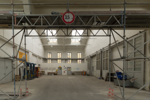 Lagerhalle mit ausgedehnten Gerüsten, Verkehrskegeln, Metallstangen, einem Schild, Maschinen, einer Tür, Fenstern, Wänden, Deckenleuchten und einem Dach.