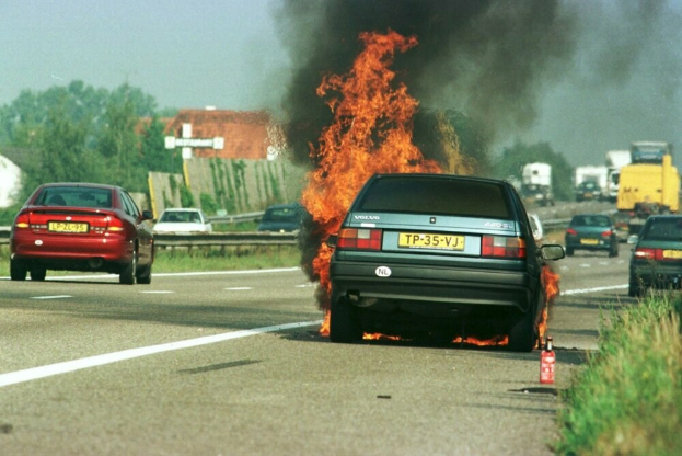 Ein Auto, das in Flammen steht, auf der Seite der Straße, umgeben von anderen Fahrzeugen, mit Bäumen, Gebäuden und einem klaren blauen Himmel im Hintergrund; Gras und ein Feuerlöscher sind auf der rechten Seite zu sehen.