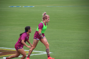 Zwei Frauen beim Fußballspielen auf einem Feld, eine trägt ein grünes T-Shirt und Shorts, die andere ein pinkes Kleid, beide tragen Sportkleidung und haben Tätowierungen an Händen und Füßen.