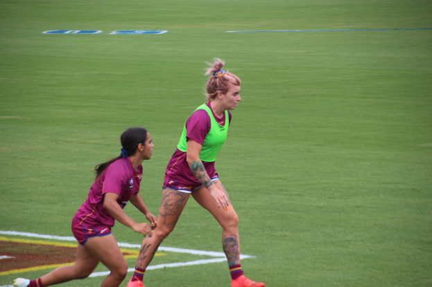 Zwei Frauen beim Fußballspielen auf einem Feld, eine trägt ein grünes T-Shirt und Shorts, die andere ein pinkes Kleid, beide tragen Sportkleidung und haben Tätowierungen an Händen und Füßen.