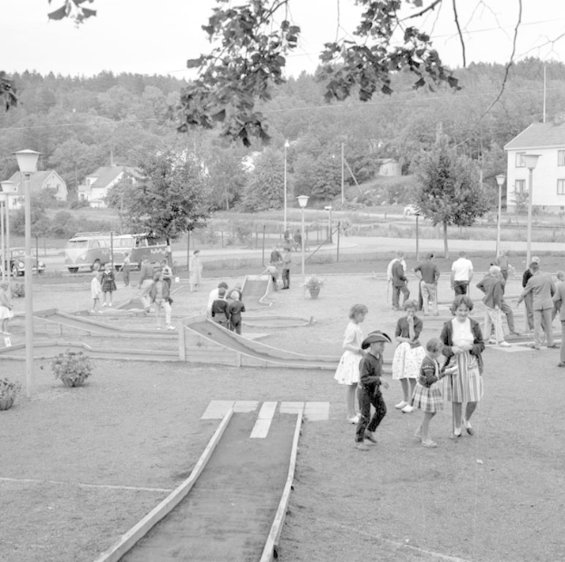 Schwarze und weiße Aufnahme von Menschen, die Minigolf in einem Park mit üppiger Vegetation, Laternenmasten, geparkten Fahrzeugen und Gebäuden im Hintergrund spielen.