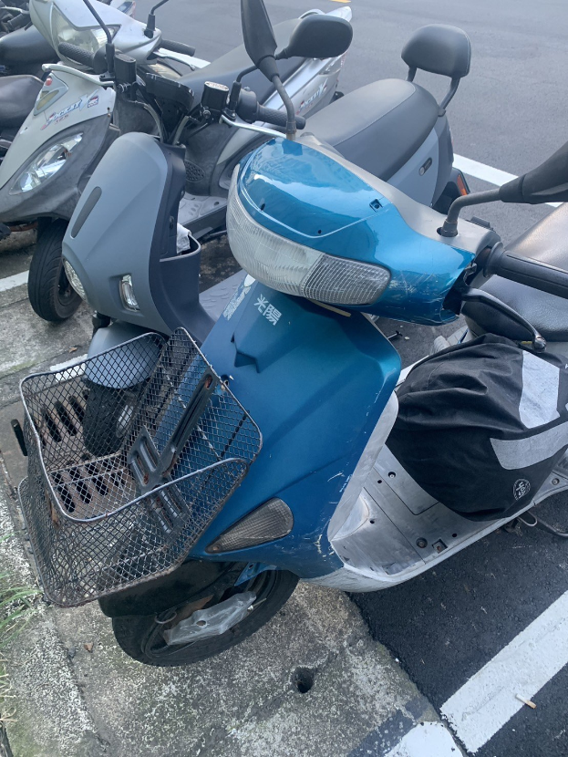 Ein blaues Rollerfahrrad in einem Parkplatz neben anderen Motorrädern geparkt, mit Gras auf der linken Seite.