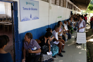 Eine Gruppe von Menschen sitzt auf Bänken vor einem Gebäude mit einer Texttafel an der Wand, einer Tür links und Bäumen im Hintergrund und wartet an einer Wahllokal in Guadalajara, Mexiko.