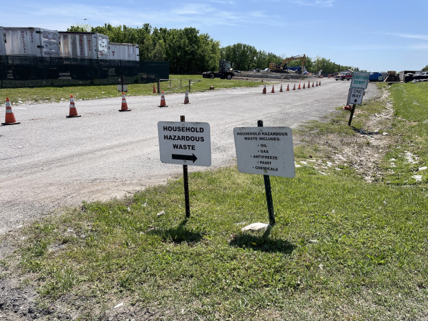 Ein Straßenschild mit der Aufschrift "Haushaltsgefährlicher Abfall" mit Gras, Verkehrskegeln, Fahrzeugen, Bäumen und einem bewölkten Himmel im Hintergrund.