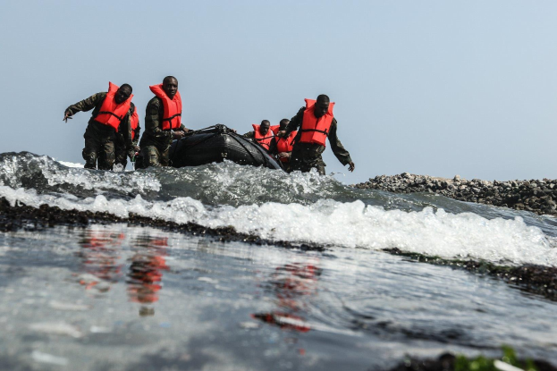 Eine Gruppe von Menschen in Schwimmwesten auf einem Boot im Wasser, umgeben von Felsen auf der rechten Seite und einem klaren Himmel, wahrscheinlich Teil eines Suchtrupps nach Uberlebenden.