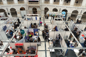 Eine große Gruppe von Menschen versammelte sich in einem Raum voller Stände, Banner, Tische, Stühle und Ballons, mit einem Gebäude mit Bögen, Säulen, Geländern und einem Bildschirm im Hintergrund.