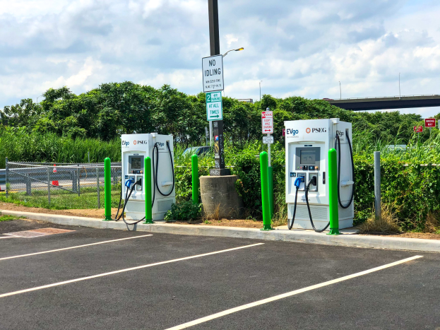 Elektrofahrzeug-Ladestation auf einem Parkplatz mit umgeben-der Infrastruktur, Grünfläche und einer Brücke im Hintergrund unter einem bewölkten Himmel.