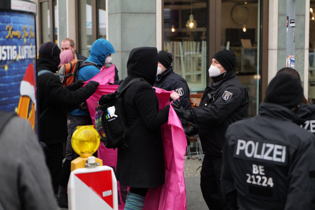 Eine Gruppe von Menschen vor einem Glasgebäude in Berlin, mit einem Polizeibeamten, der eine Frau festnimmt; einige halten Kleidung, andere tragen Mützen und Taschen, und ein Pfahl mit Text ist links sichtbar.