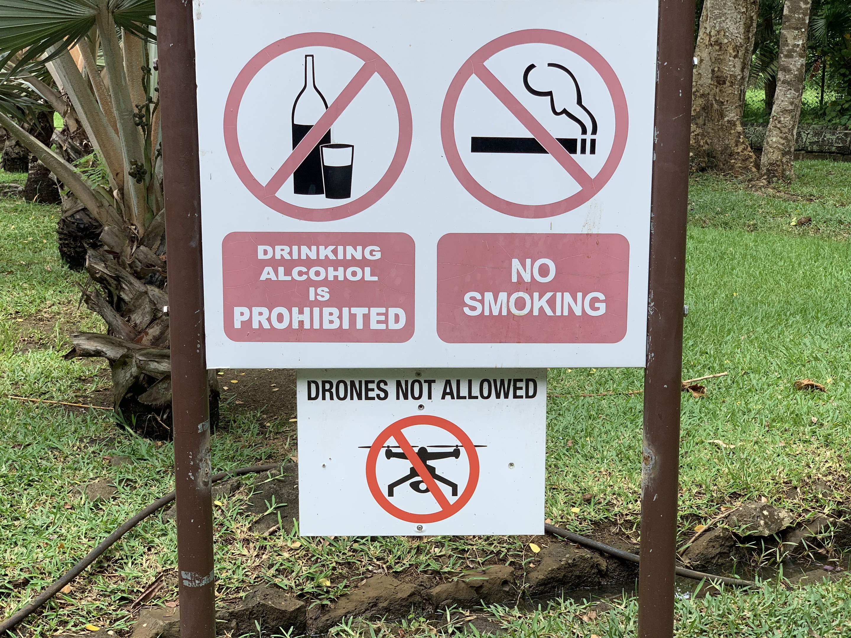 Schild am Straßenrand, das das Trinken von Alkohol und Rauchen verbietet, mit Bäumen und Gras im Hintergrund.