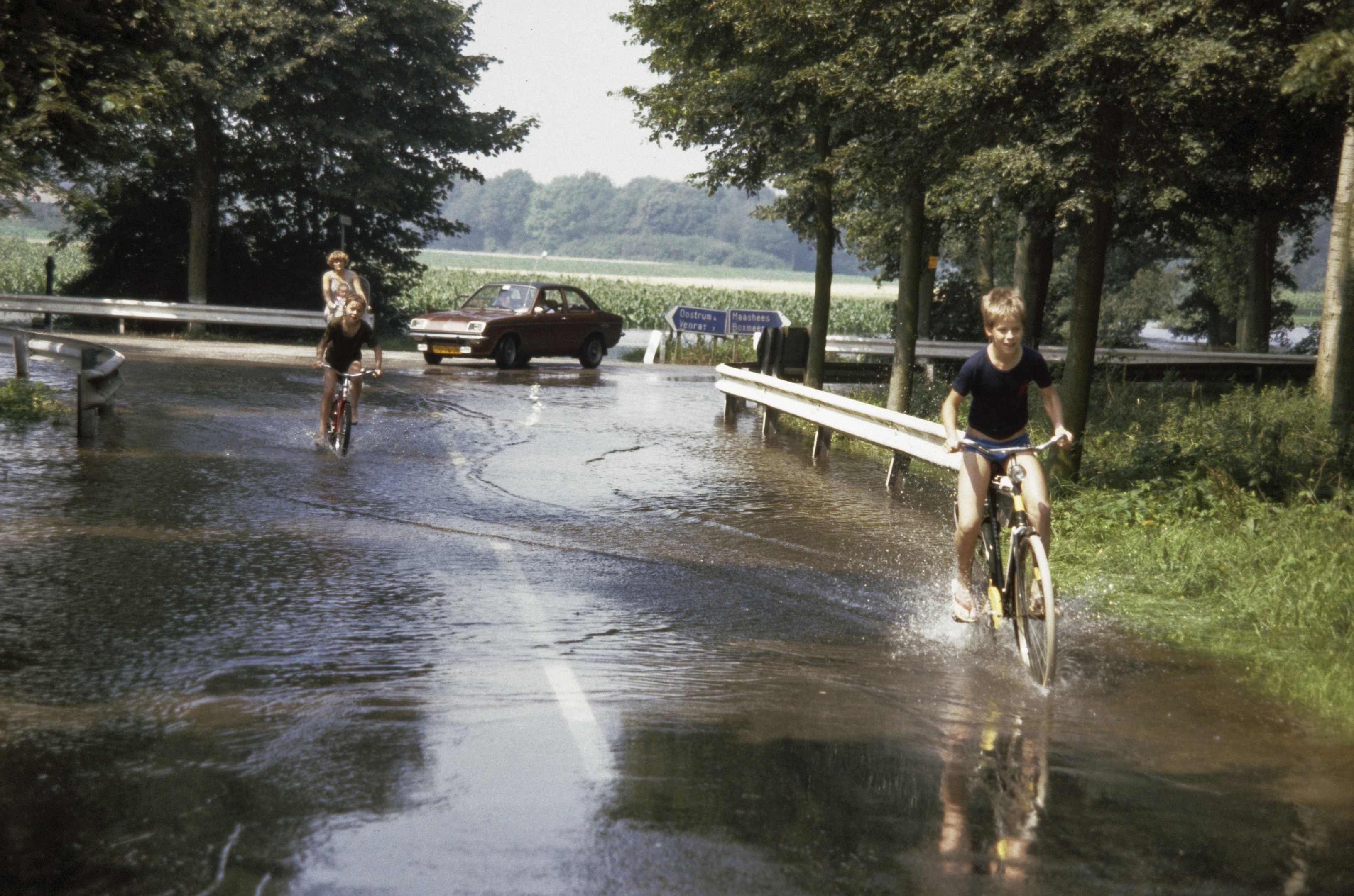 Eine Gruppe von Menschen, die auf Fahrrädern eine überschwemmte Straße entlangfahren, mit einem danebenfahrenden Auto, gesäumt von Geländern und Bäumen unter einem klaren blauen Himmel.