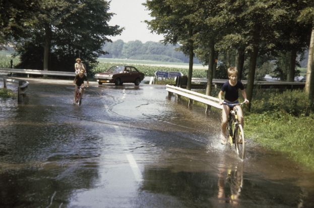 Eine Gruppe von Menschen, die auf Fahrrädern eine überschwemmte Straße entlangfahren, mit einem danebenfahrenden Auto, gesäumt von Geländern und Bäumen unter einem klaren blauen Himmel.